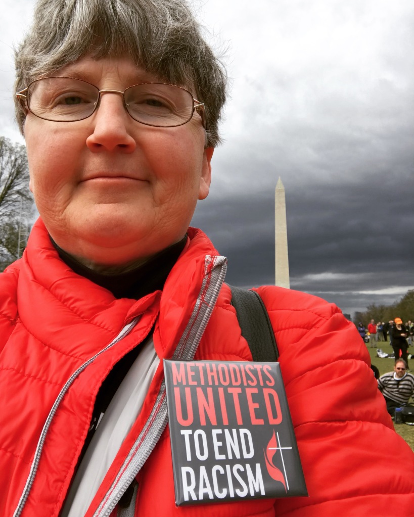 Woman in red coat, with a pin that says "Methodists United to End Racism". A stormy sky and the Washington Monument are behind her.